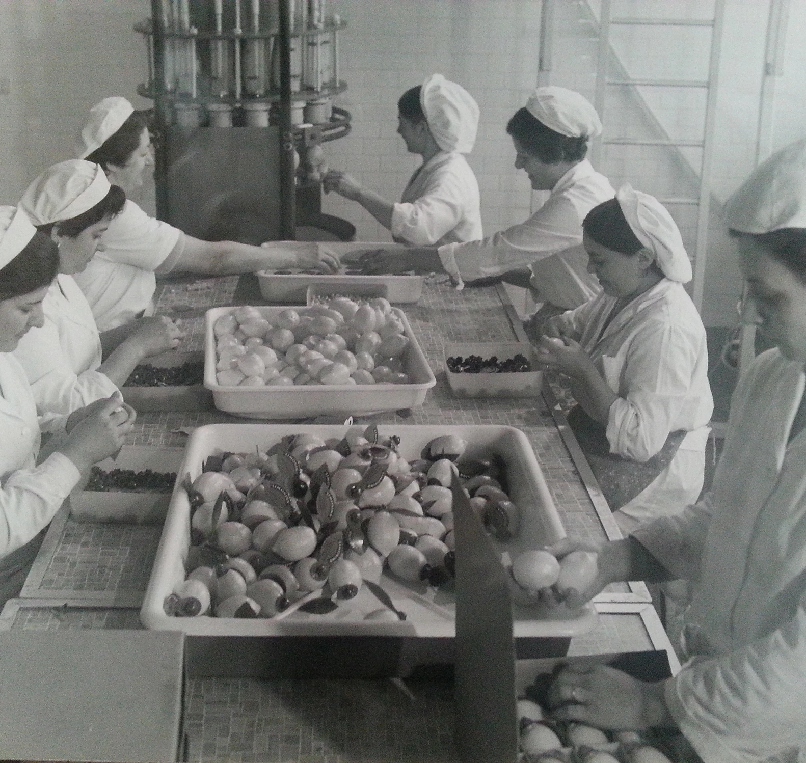 Sei donne in divisa e con il berretto sono al lavoro a un tavolo, intente a selezionare e confezionare caramelle o cioccolatini in scatole, in un ambiente industriale con vassoi pieni di dolciumi incartati.
