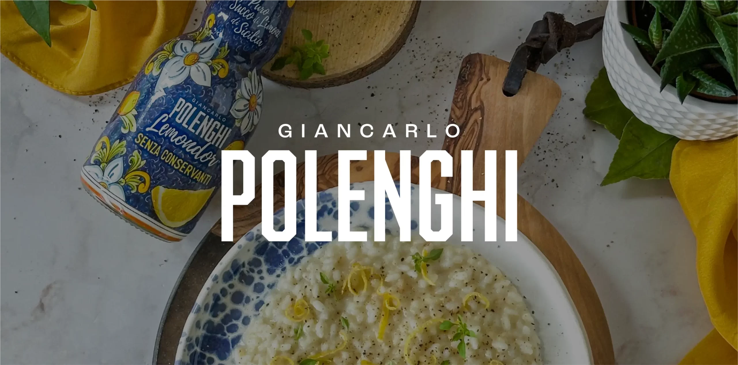 Caraffina Polenghi di succo di limone decorata, accanto a un risotto al limone guarnito con scorza, tagliere e pianta su tavola.
