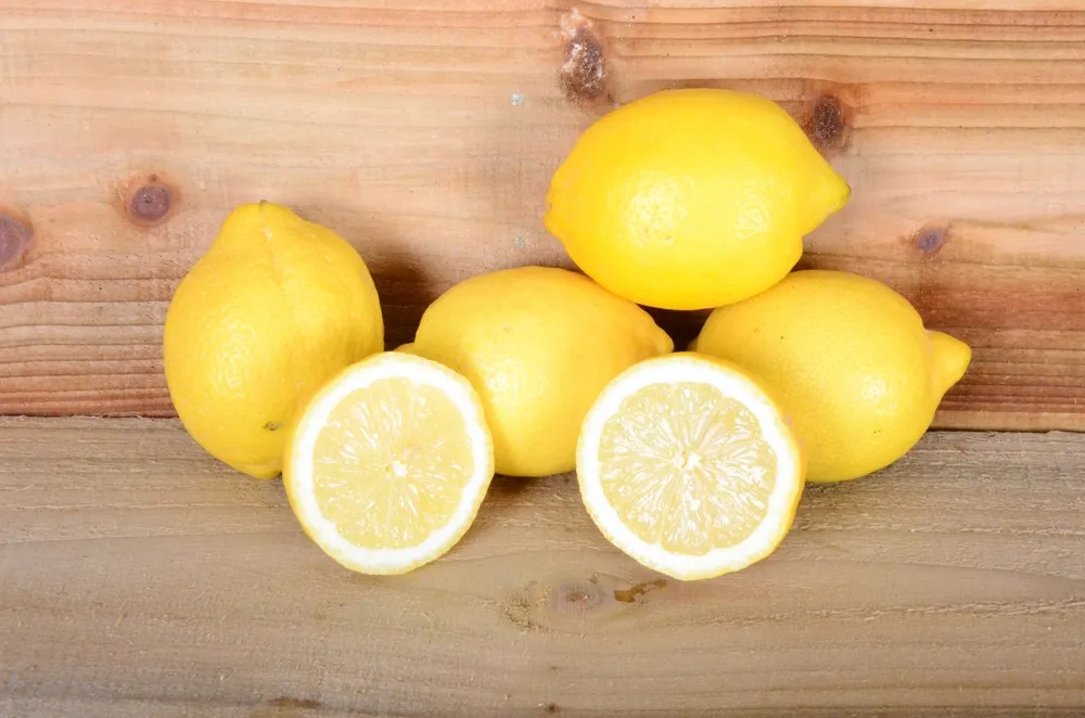 Five Sorrento lemons arranged on a light wooden surface, with two sliced in half in the foreground to show the pale pulp and medium-thick rind.