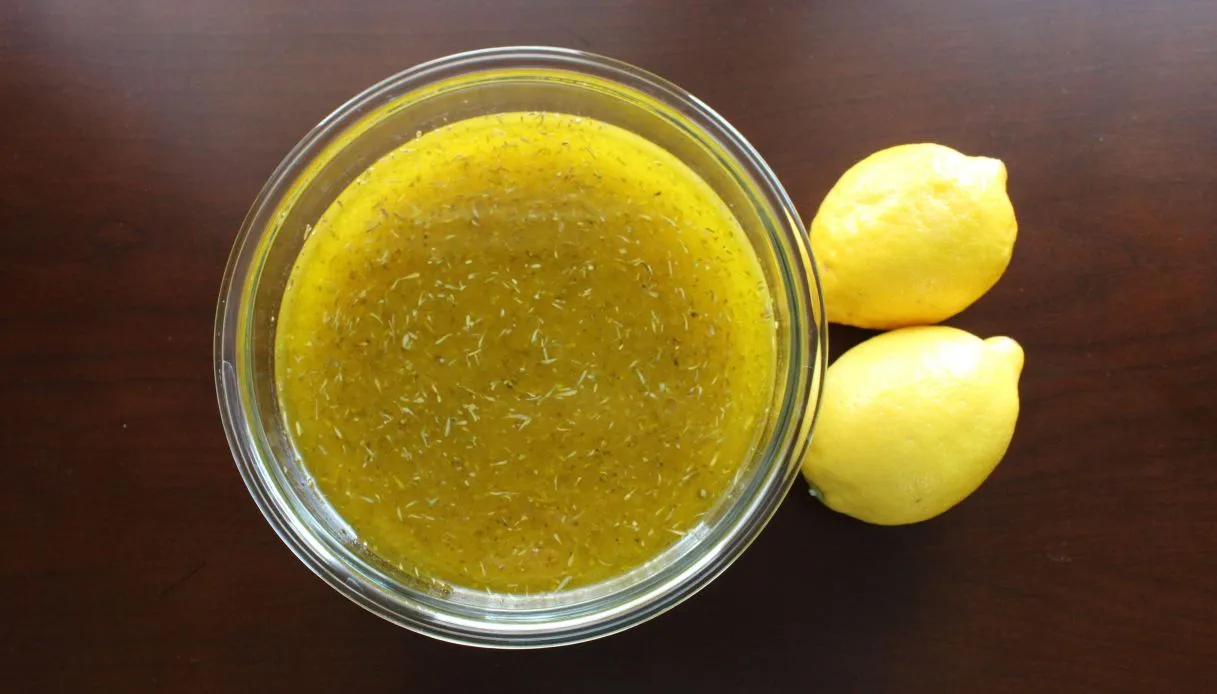 Una ciotola di vetro contenente un condimento per insalata giallo con erbe aromatiche, accanto a due limoni interi, il tutto su una superficie marrone scuro.