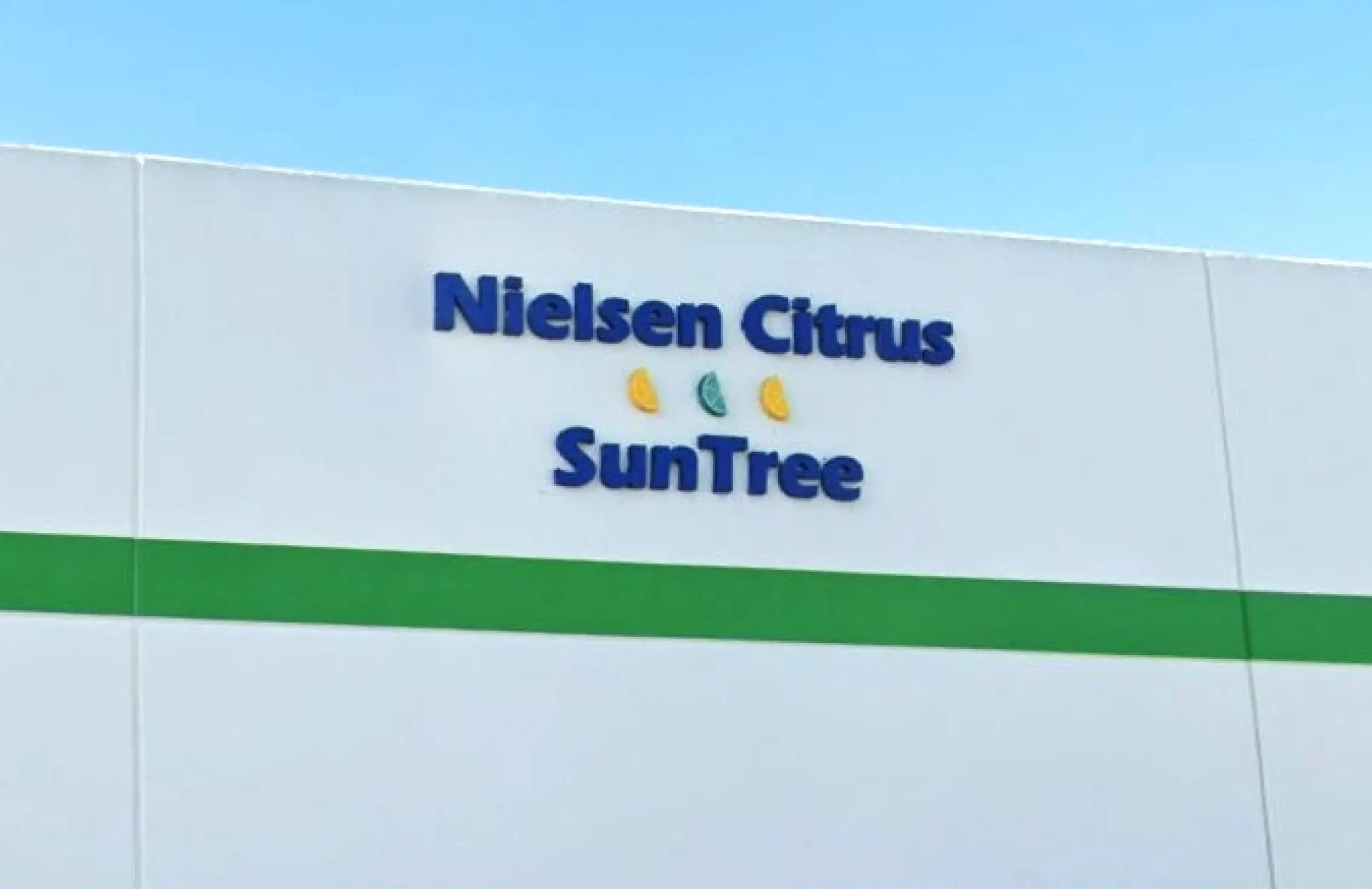 L'esterno di un edificio bianco con l'insegna “Nielsen Citrus SunTree” e tre piccole icone raffiguranti agrumi tra le parole, sotto un cielo azzurro e limpido.