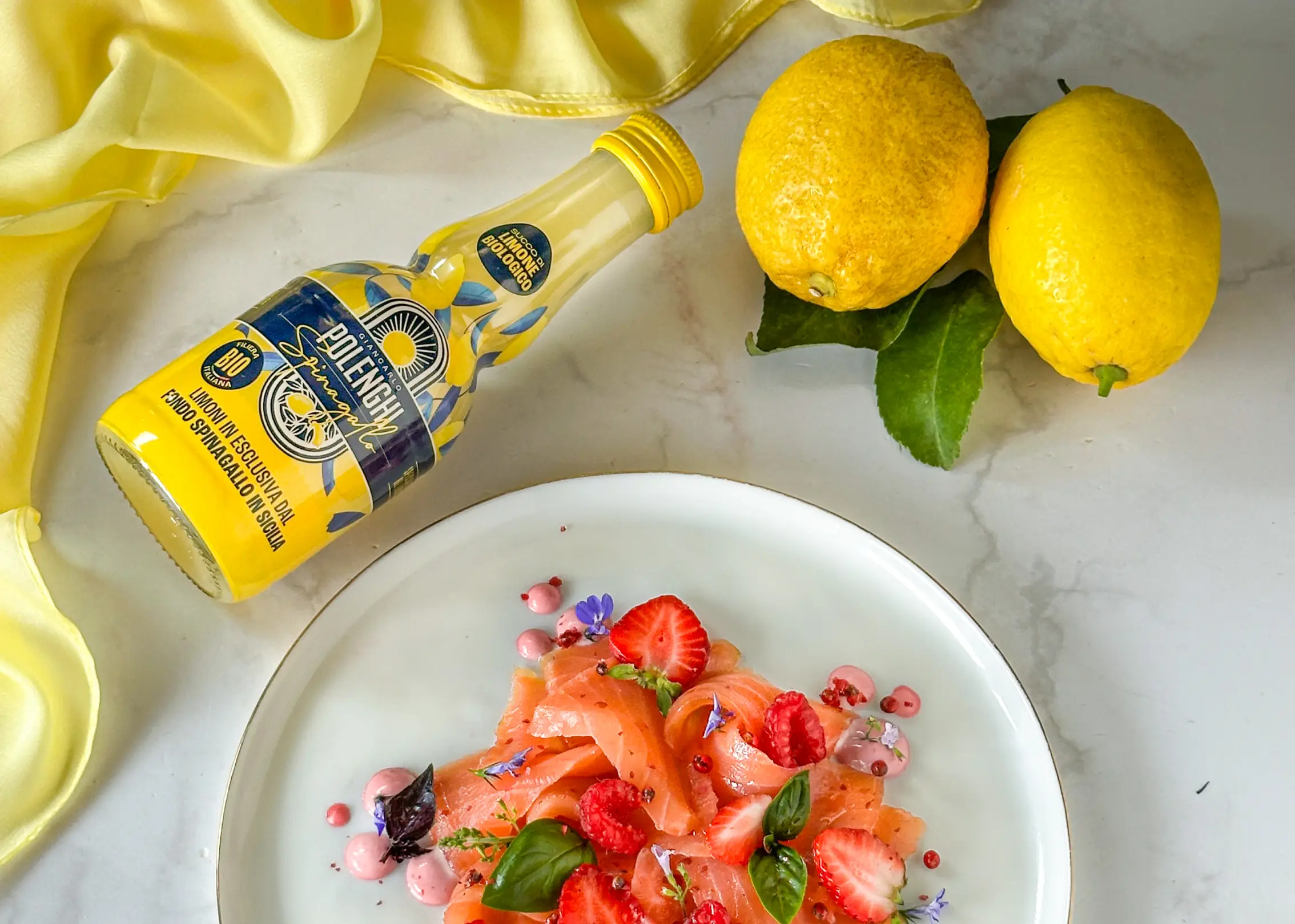 A plate of smoked salmon with strawberries, raspberries, and edible flowers sits near two lemons and a bottle of yellow lemon dressing on a marble surface with a yellow cloth.
