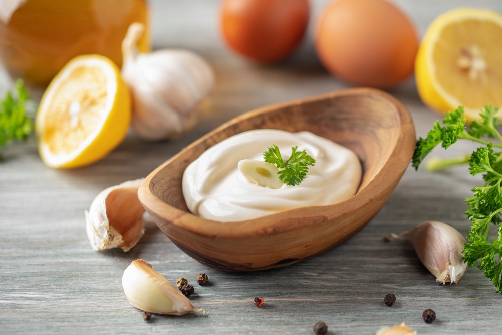 Una ciotola di legno contenente salsa aioli cremosa guarnita con prezzemolo e aglio, circondata da spicchi d’aglio, metà di limone, uova ed erbe aromatiche su un piano di legno.