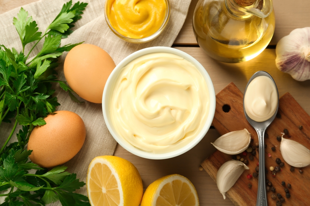 Una ciotola di maionese circondata da uova, prezzemolo, metà di limone, aglio, un barattolo di olio, senape e un cucchiaio con della maionese su un tagliere di legno con grani di pepe.