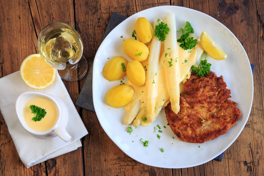 Un piatto con una cotoletta impanata, asparagi bianchi con salsa olandese, patate lesse, spicchi di limone e prezzemolo. Accanto al piatto, su un tavolo rustico di legno, ci sono un bicchiere di vino bianco, mezzo limone e della salsa extra in una tazzina.