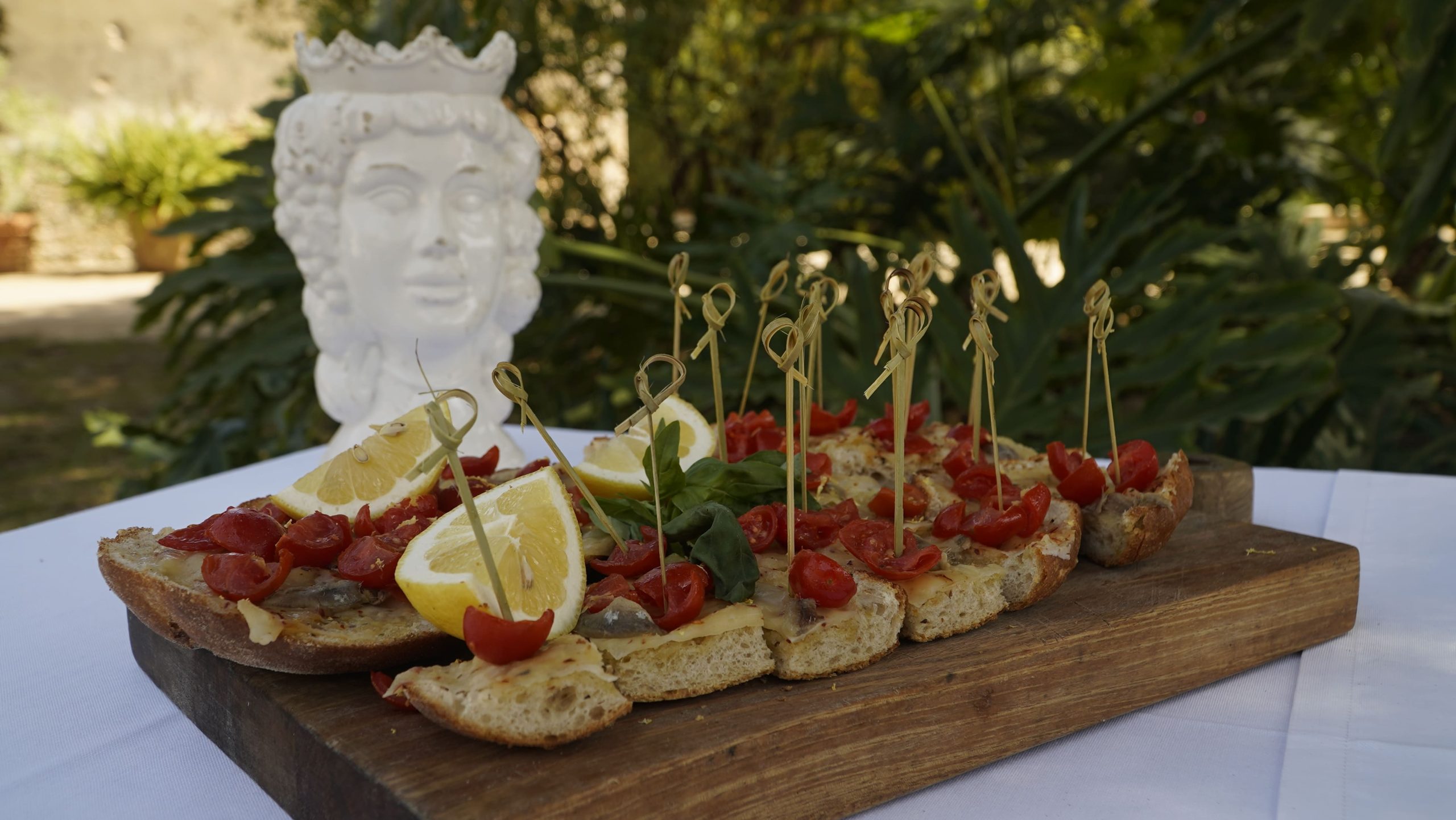 A wooden cutting board with several slices of bruschetta arranged on it, garnished with cherry tomatoes, lemon wedges, and bamboo toothpicks, with a decorative ceramic bust in the background.