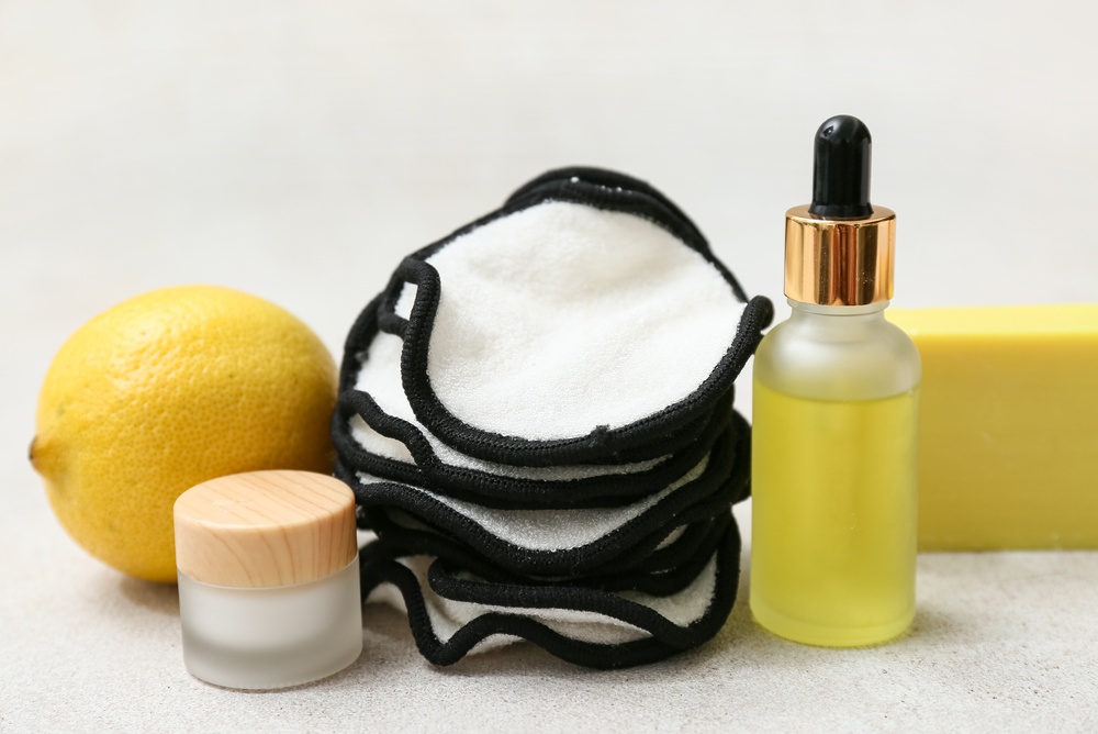 Una pila di dischetti struccanti riutilizzabili accanto a un limone, un vasetto con un coperchio di legno, un flacone di vetro con contagocce contenente un olio giallo e una saponetta gialla.