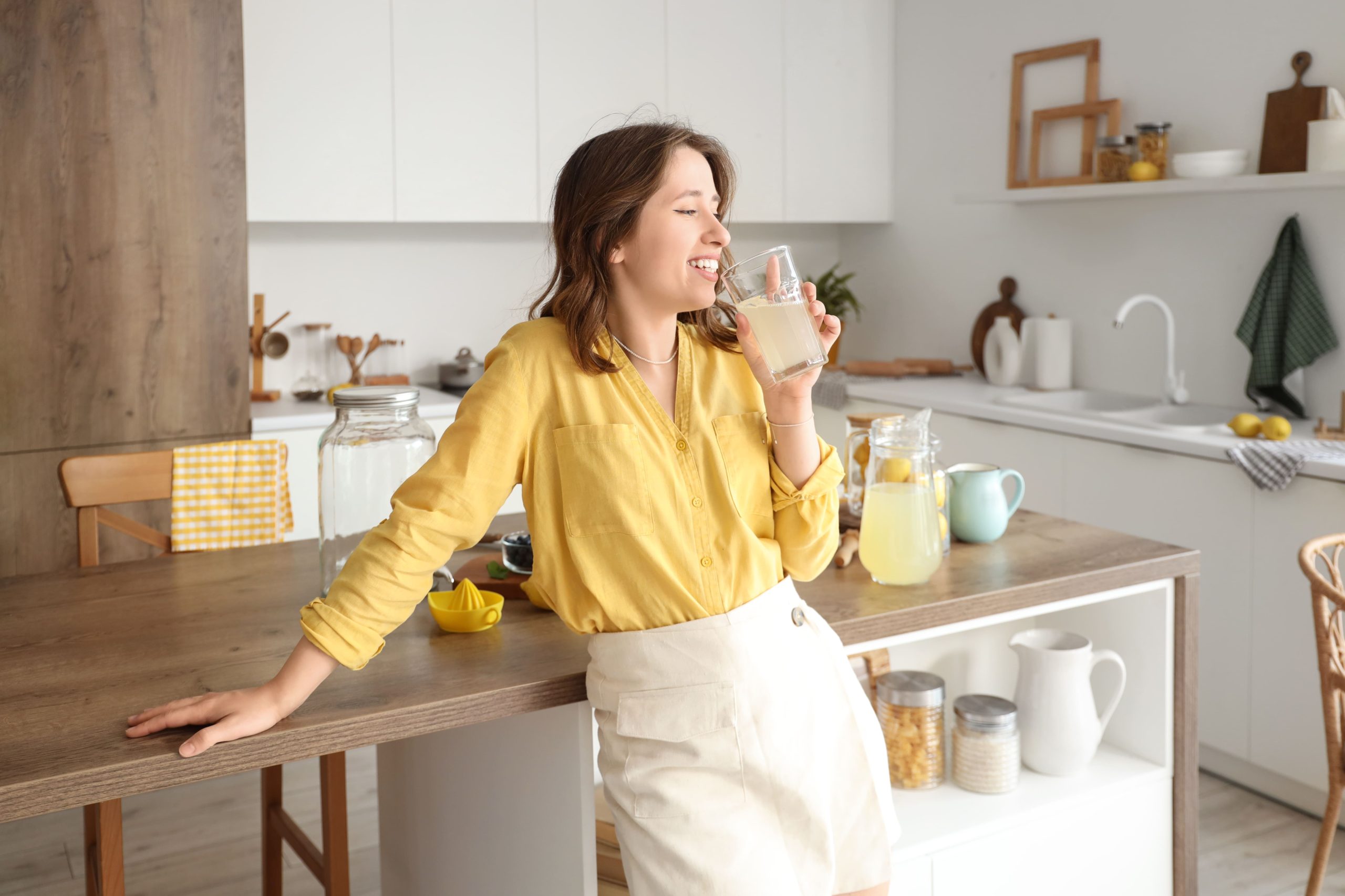 Una donna con una maglietta gialla che sorride mentre beve un bicchiere di limonata in cucina.
